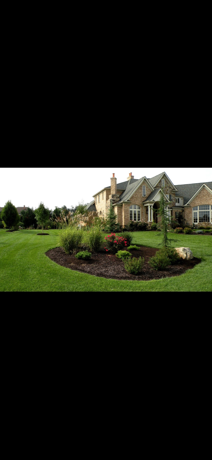 Custom mulch bed island with ornamental grasses and roses framing a stone-and-brick estate home